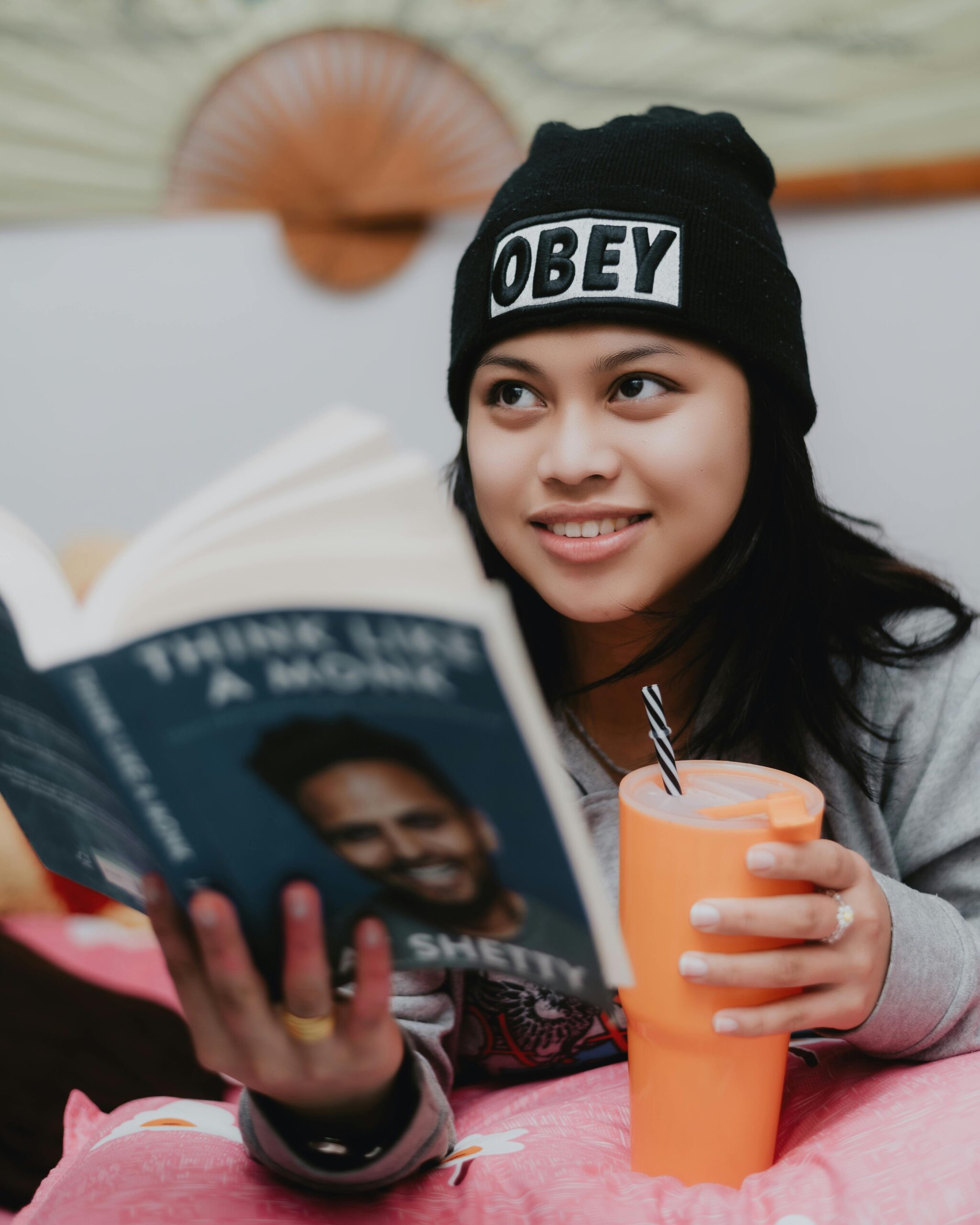 Smiling woman in beanie reading 'Think Like a Monk' while holding a cup indoors.